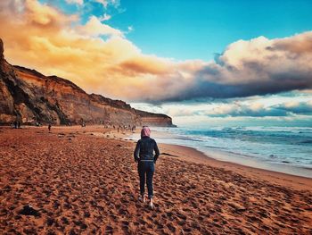 Rear view of man walking at sandy beach against sky during sunset