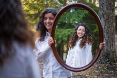 Smiling young woman holding mirror for sister in forest