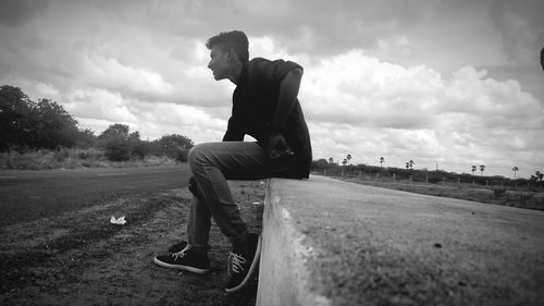 Side view of smiling young man sitting on concrete wall against sky