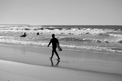 Surfer walking with surfboard on shore at beach against clear sky