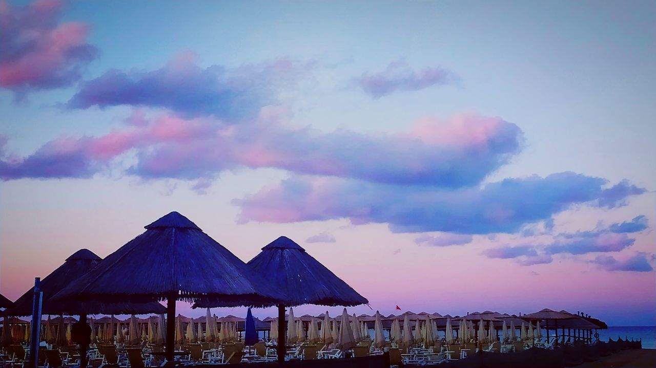 sky, cloud, beach, dusk, evening, water, nature, sunset, beauty in nature, architecture, land, sea, horizon, hut, scenics - nature, travel destinations, thatched roof, tranquility, umbrella, built structure, outdoors, no people, beach hut, tranquil scene, ocean, idyllic, vacation, trip, parasol, travel, holiday, summer, roof, building exterior, environment, tourism, landscape, blue, sunshade