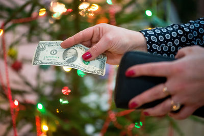 Cropped hands of woman holding paper currency against christmas tree