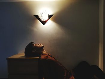 Man sleeping while sitting by illuminated lighting equipment on wall at home