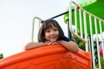 Low angle portrait of smiling girl sitting on slide against sky in playground
