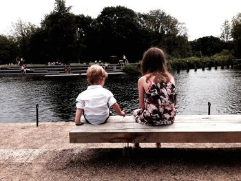 Rear view of siblings sitting on bench at riverbank