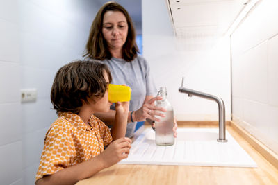 Unrecognizable kid in ornamental clothes drinking water from glass near woman with bottle at home