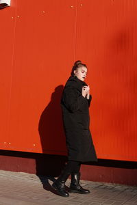 Fashionable teenage girl wearing black warm clothing standing against red wall during sunny day