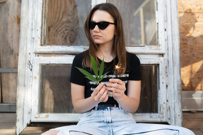 Young woman wearing sunglasses holding leaf and sparkler while sitting against window