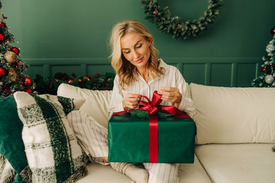 A young woman in pajamas is sitting on the couch unwrapping a christmas present.