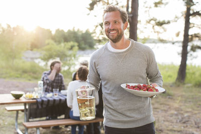 Happy man holding beer jug and strawberries with friends sitting at picnic table in background