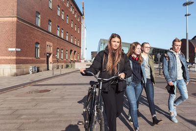 Teenage girl holding bicycle while walking with friends on footpath in city against clear sky