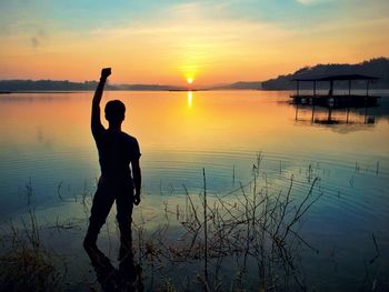 Silhouette man with hand raised standing in sea against sky during sunset