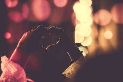 Cropped image of woman forming heart shape against lens flare at night