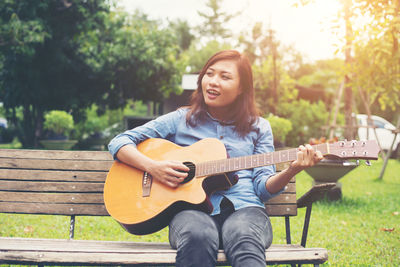 Young woman playing guitar while sitting on bench in park