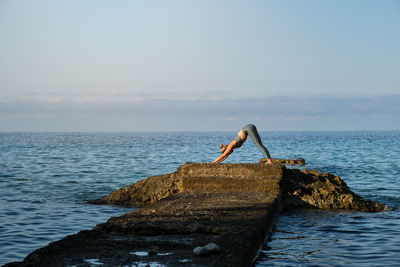 Side view of distant unrecognizable woman standing in downward facing dog position while practicing yoga on rocky pier at seaside