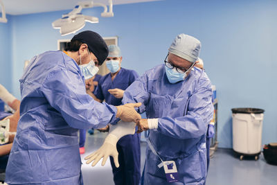 Assistant helping doctor to wear protective glove while standing in operation room