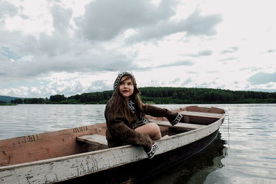 Girl looking away while sitting in rowboat on lake against cloudy sky