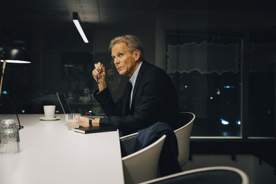 Side view of thoughtful senior male professional looking away while sitting with laptop at illuminated desk working late