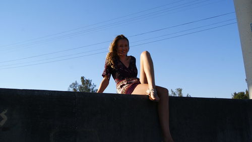 Low angle portrait of smiling young woman sitting on retaining wall against clear blue sky during sunny day