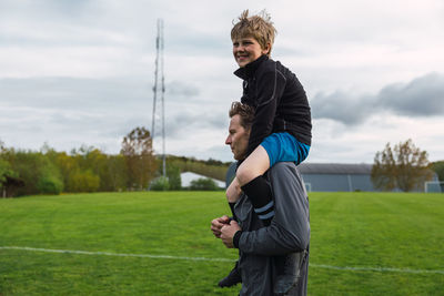 Cheerful father carrying teenage boy on shoulder while standing on football field