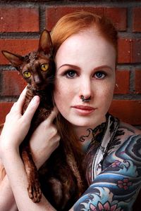 Portrait of redhead woman holding cat against brick wall