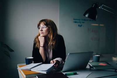 Thoughtful young businesswoman sitting with document and laptop at creative office