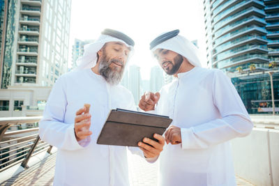 Men wearing dish dash discussing over digital tablet on footbridge in city