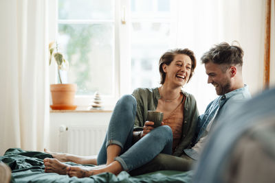 Happy woman with mug sitting with man on bed at home