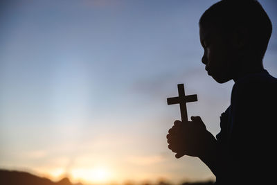 Silhouette boy holding cross against sky during sunset