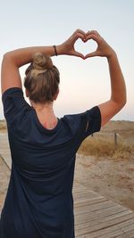 Rear view of mid adult woman with arms raised making heart shape while standing against clear sky