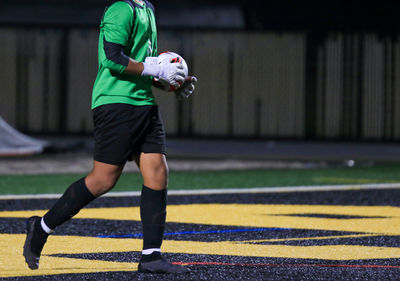 A goalkeeper in green jersey holds a soccer ball while walking on the field during a nighttime game.