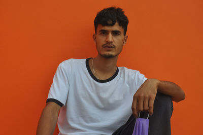 A young guy looking at camera with holding face mask, sitting against orange wall with copy space 
