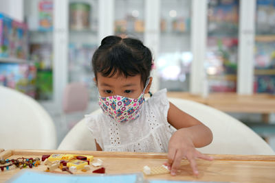 Cute little girl with protective face mask playing plastic building blocks at table.