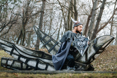 Man in costume looking away while sitting against bare trees in forest