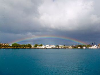 Sea in front of city against rainbow in cloudy sky