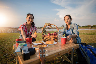 Portrait of smiling friends having food and drink while sitting against sky at sunset