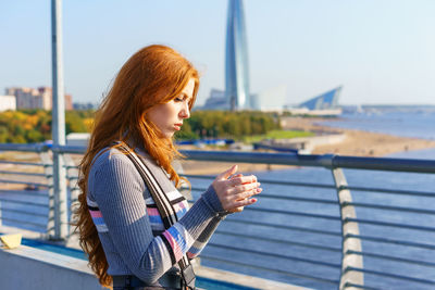 Young red-haired woman with phone in her hand stands on bridge overlooking city