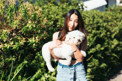 Portrait of smiling young woman carrying white dog against plants during sunny day
