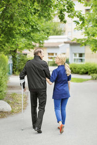 Full length rear view of female caretaker walking with disabled senior man on street