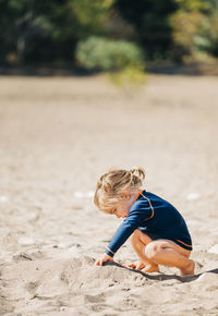 Side view of girl playing with sand while crouching at beach during sunny day