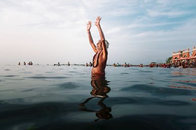 Shirtless man with arms raised standing in river against sky