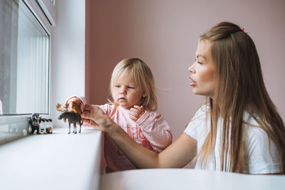 Little girl with her mother playing with animal toys on window sill in children's room at home