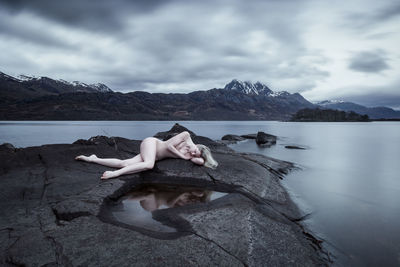 Man relaxing on rock by lake against sky