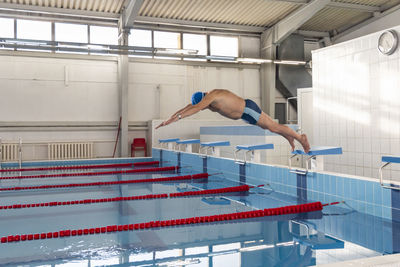 An elderly male athlete in a hat and glasses jumps from a height into the pool