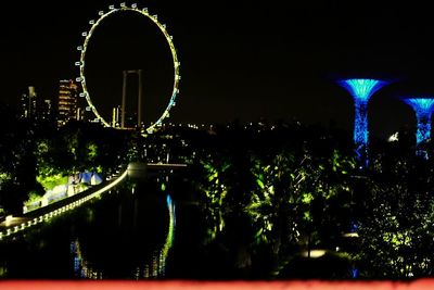 Illuminated ferris wheel by river against sky in city at night