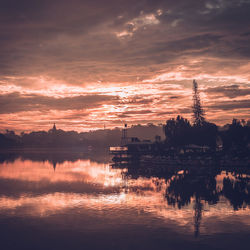 Silhouette factory by lake against sky during sunset