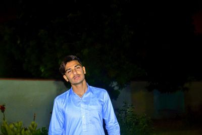 Young man looking away while standing against plants at night