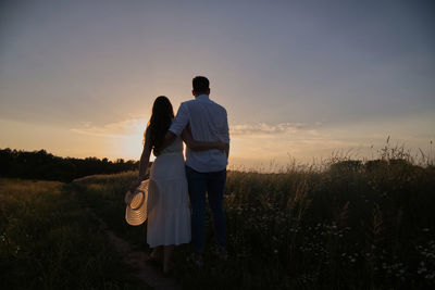 Rear view of couple standing on field against sky during sunset