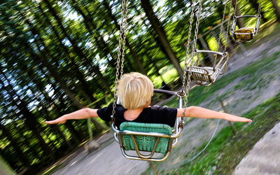 Rear view of boy on a carousel with arms outstretched against trees