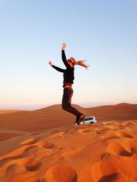 Side view of playful woman jumping at desert against clear blue sky